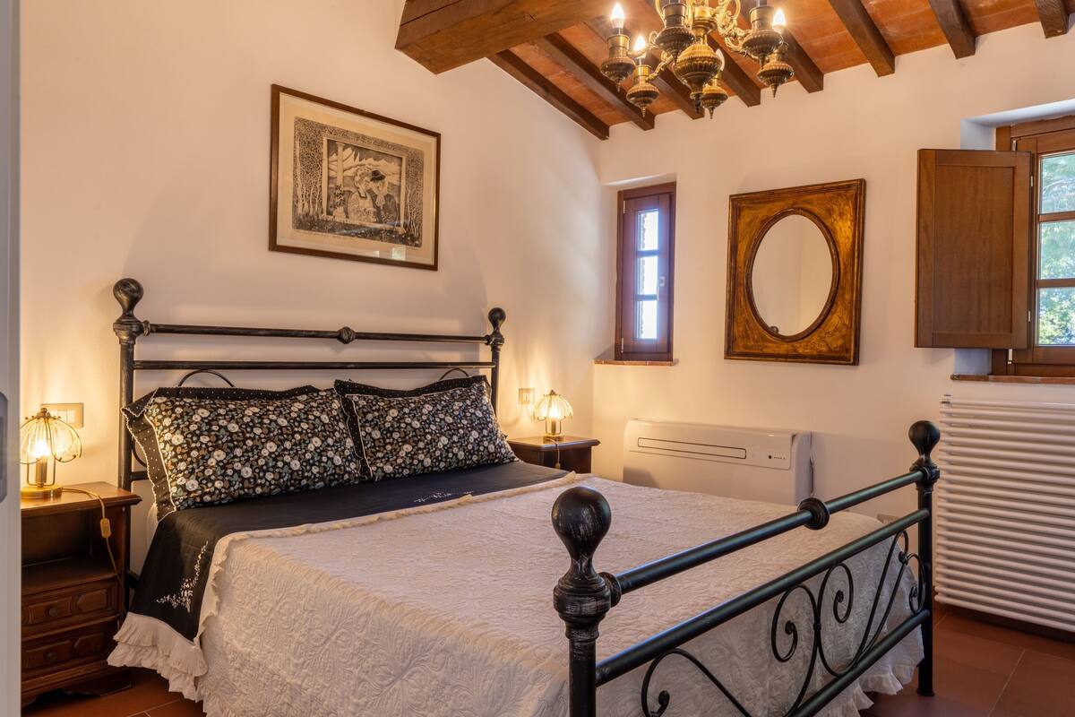 A bedroom with a bed, bedside lamps, a wooden headboard, a framed picture, a mirror, and a window with shutters.
