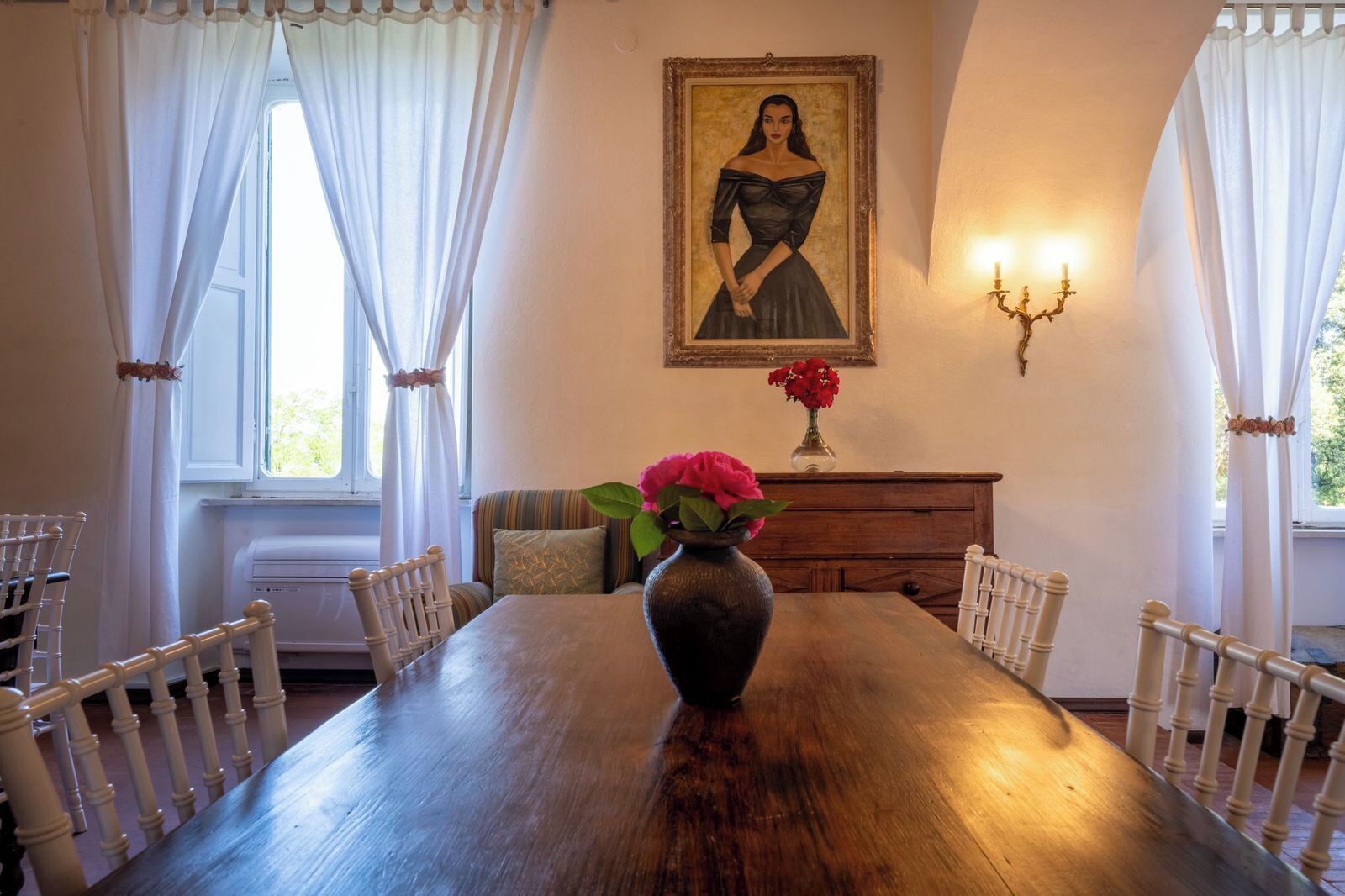 A dining room with a wooden table, white chairs, a vase with flowers, a painting on the wall, and a candelabra.