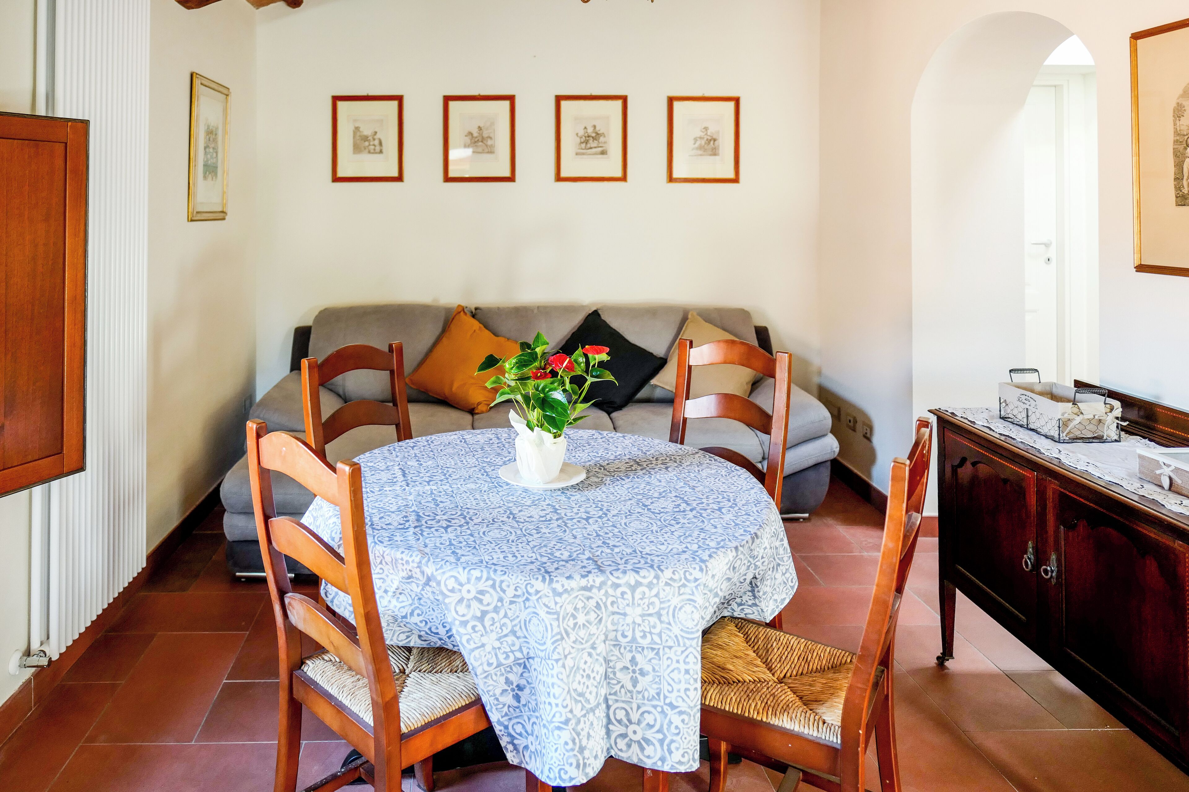 Un coin repas avec une table ronde recouverte d’une nappe bleue à motifs, entourée de chaises en bois. Un pot de fleurs avec des fleurs rouges est placé au centre.