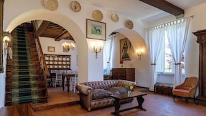 A spacious living room with a leather sofa, a wooden coffee table, and a staircase with a green carpet. There are dining tables and chairs in the background, and a chandelier hangs from the ceiling. - Tenuta dei Mandorli Pool Wifi Peccioli (Peccioli)