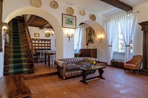 Uma sala de estar espaçosa com um sofá de couro, uma mesa de centro de madeira e uma escada com tapete verde. Mesas e cadeiras de jantar estão ao fundo, e um candelabro pendurado no teto.