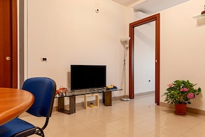 Une pièce avec un téléviseur posé sur un meuble, une chaise bleue, une petite table, une plante en pot et une porte.