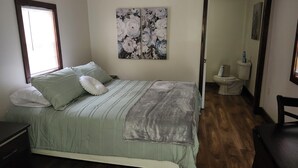 Un dormitorio con cama, ventana, un cuadro y un baño visible a través de una puerta abierta.