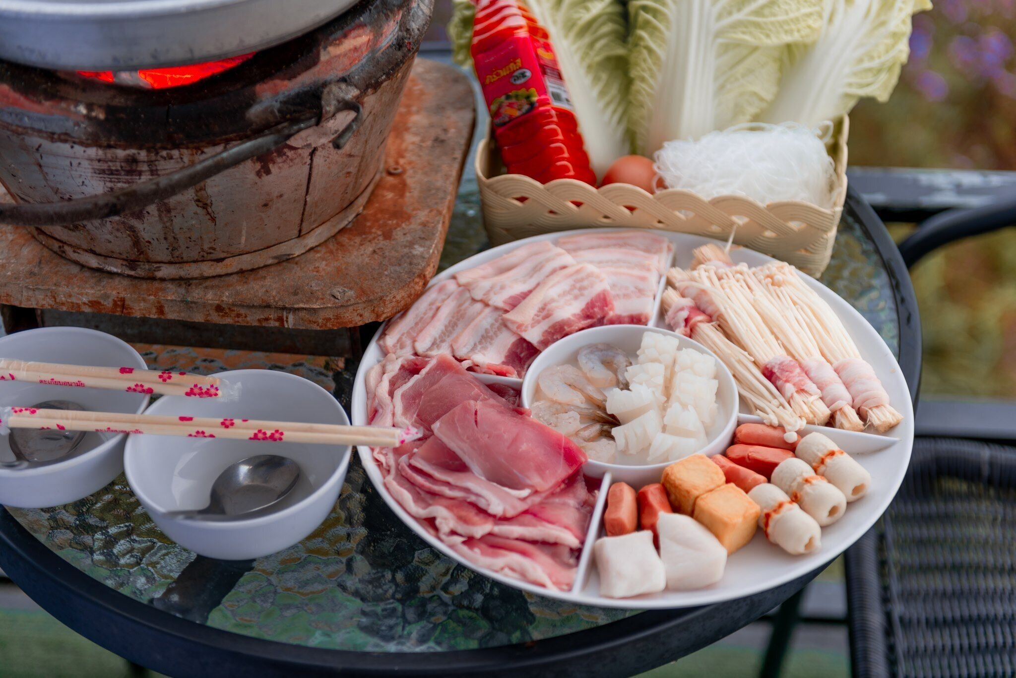 A table with various food items including meat, seafood, and vegetables, with a portable stove in the background.