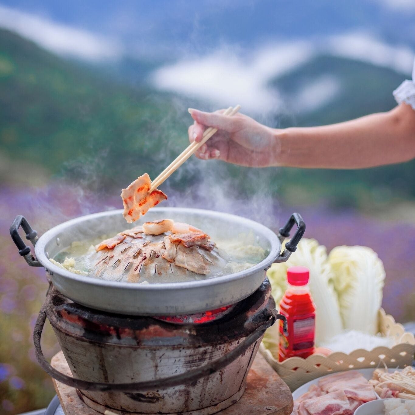 Alguém cozinha peixe numa panela sobre chama aberta, ao ar livre.