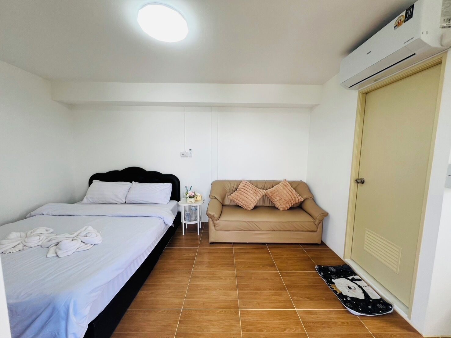 A small, clean room with a bed, a sofa, a small table with a lamp, and an air conditioning unit.