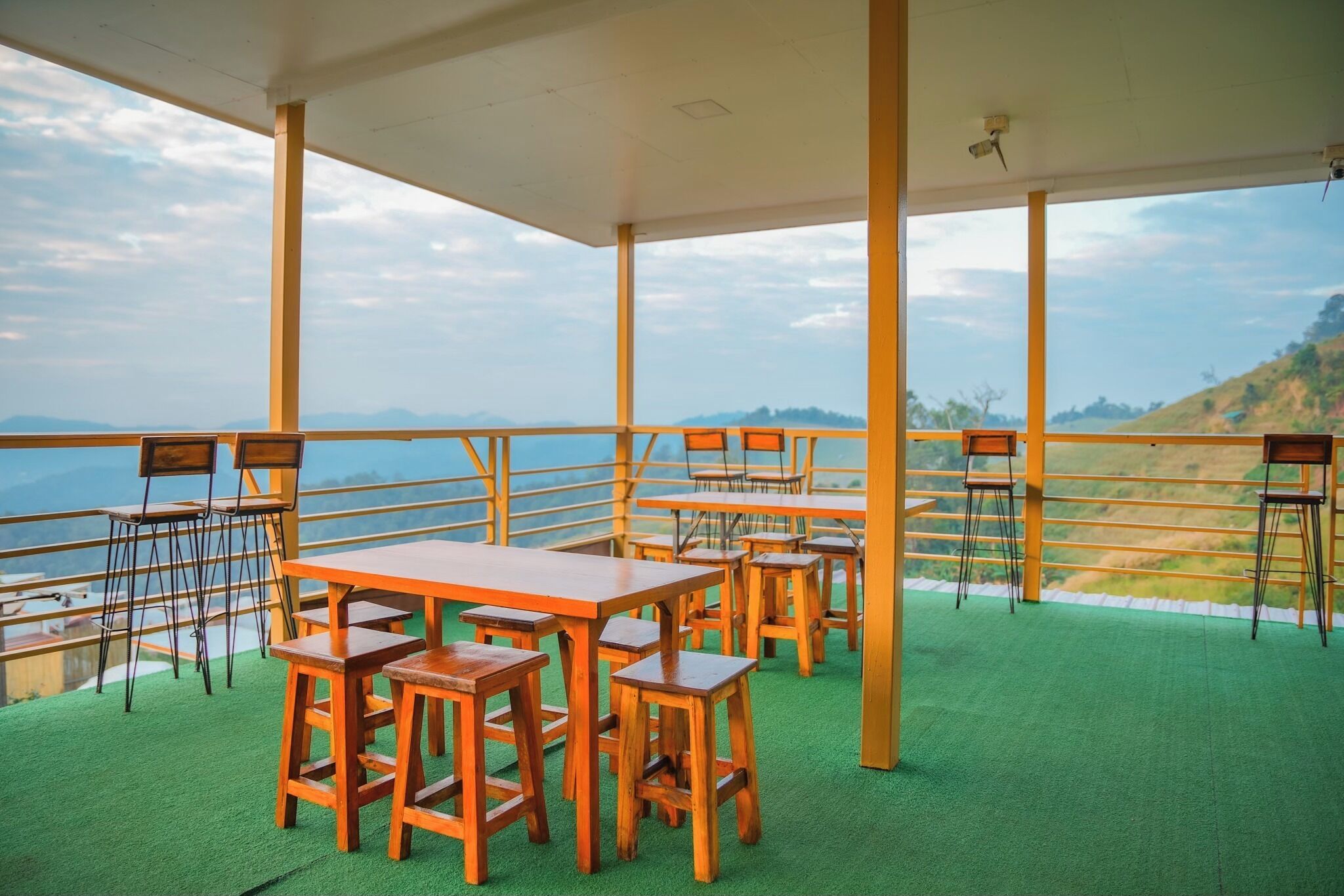 A terrace with wooden tables and chairs, a green carpet, and a railing overlooking a mountainous landscape.