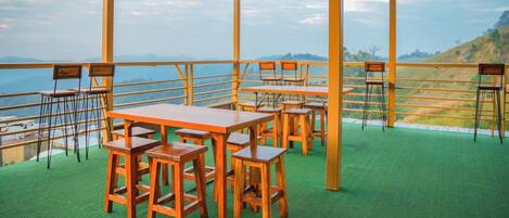 A terrace with wooden tables and chairs, a green carpet, and a railing overlooking a mountainous landscape.