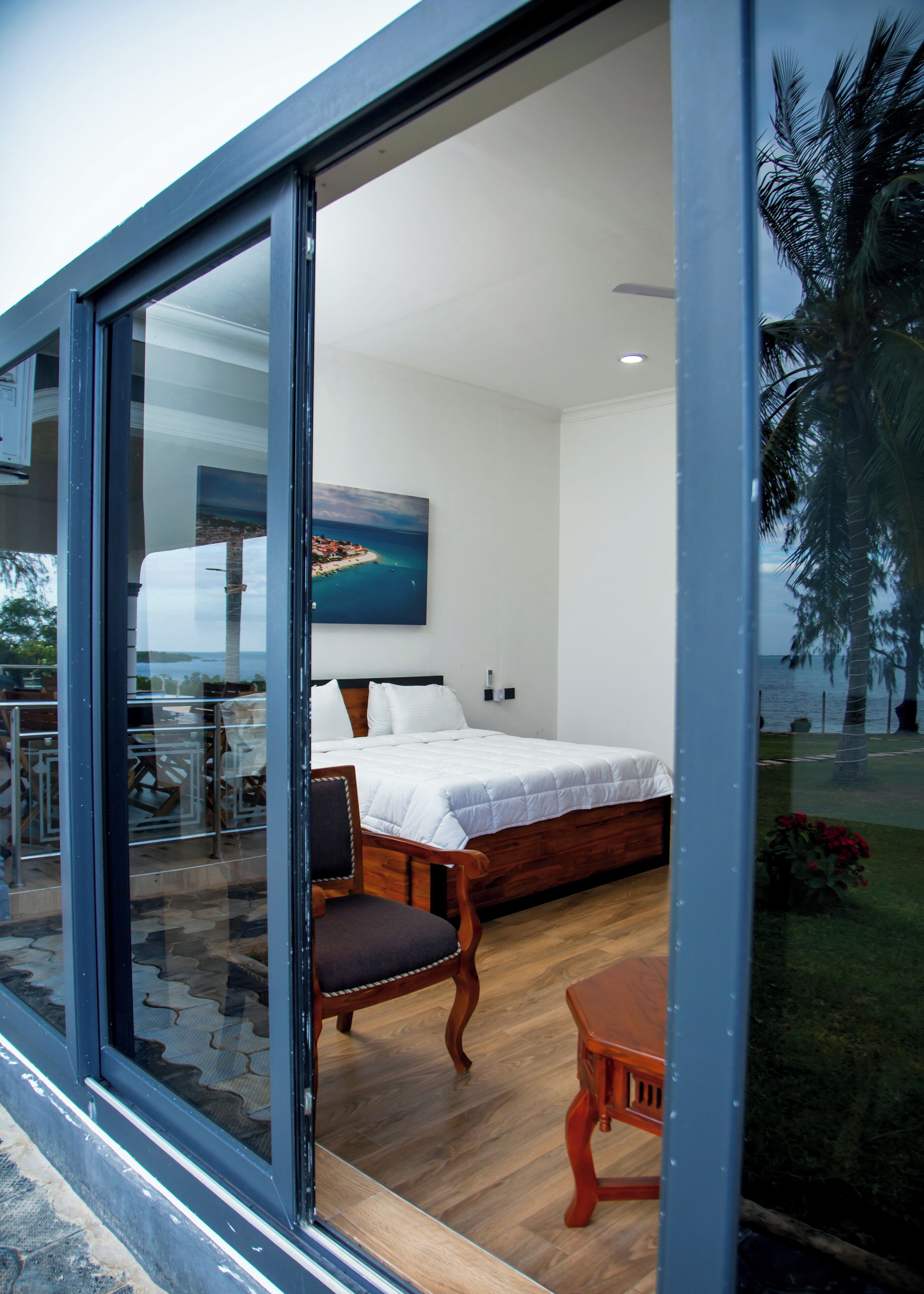 A modern bedroom with a large bed, a wooden chair, and a view of a coastal area through a glass door.