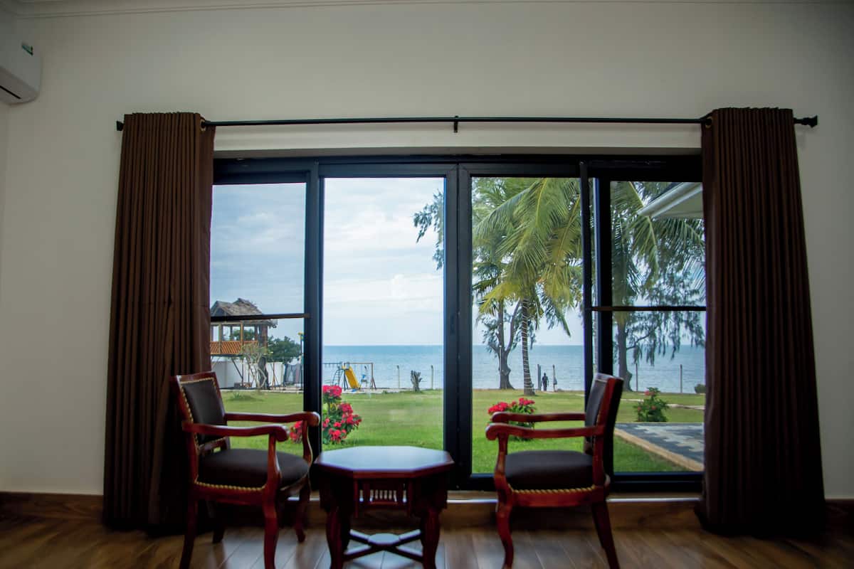 A room with a large sliding glass door leading to a balcony with a view of the sea and palm trees. There are two wooden chairs and a small table in the room.