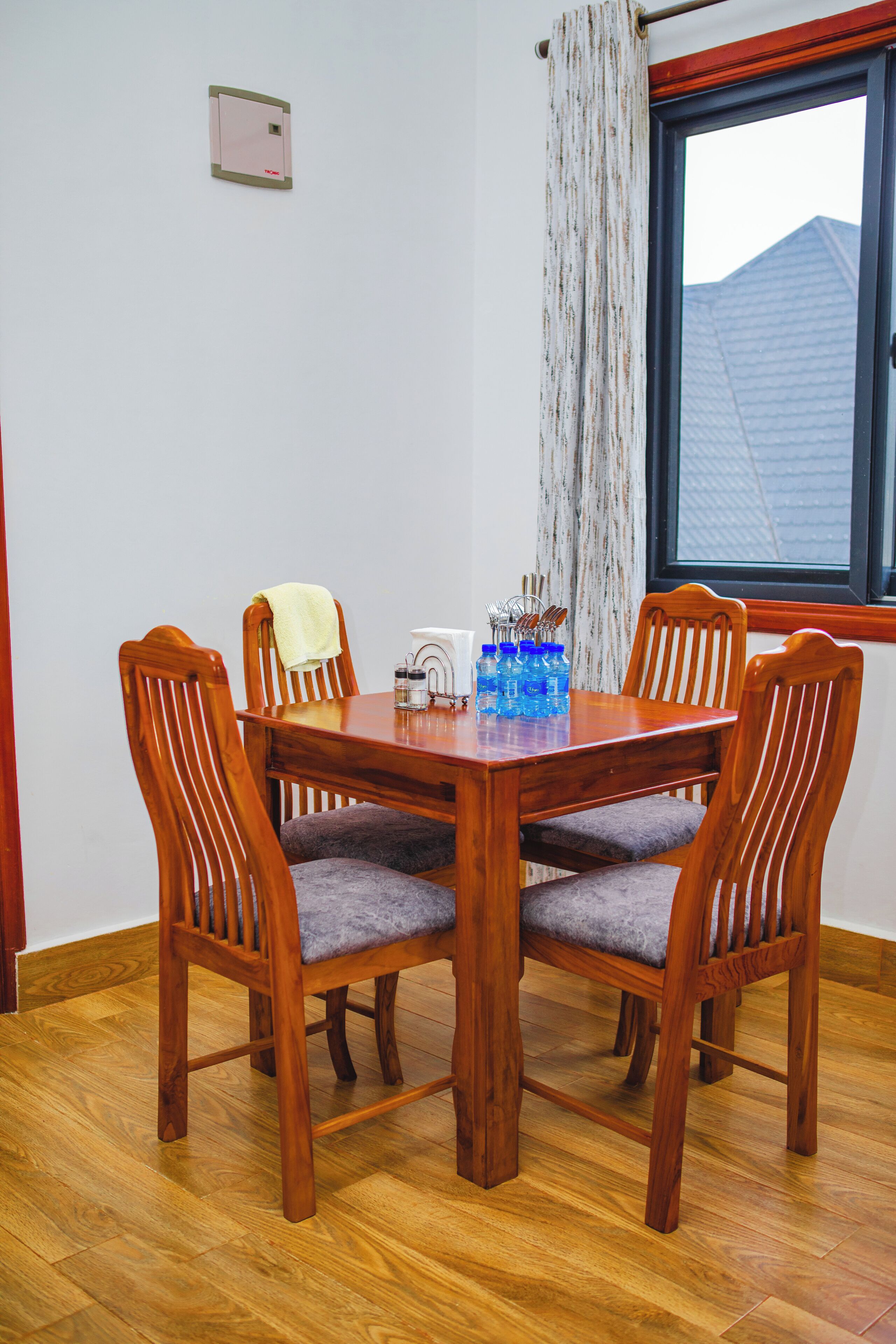 A dining area with a wooden table and four chairs, a window with a view of a building, and a light switch on the wall.
