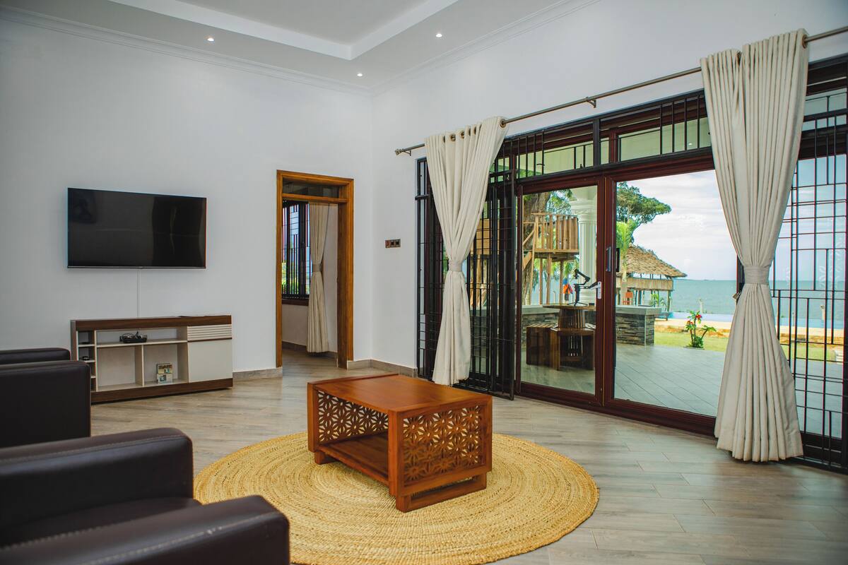A living room with a flat-screen TV, a wooden coffee table, and a view of the beach through large sliding glass doors.