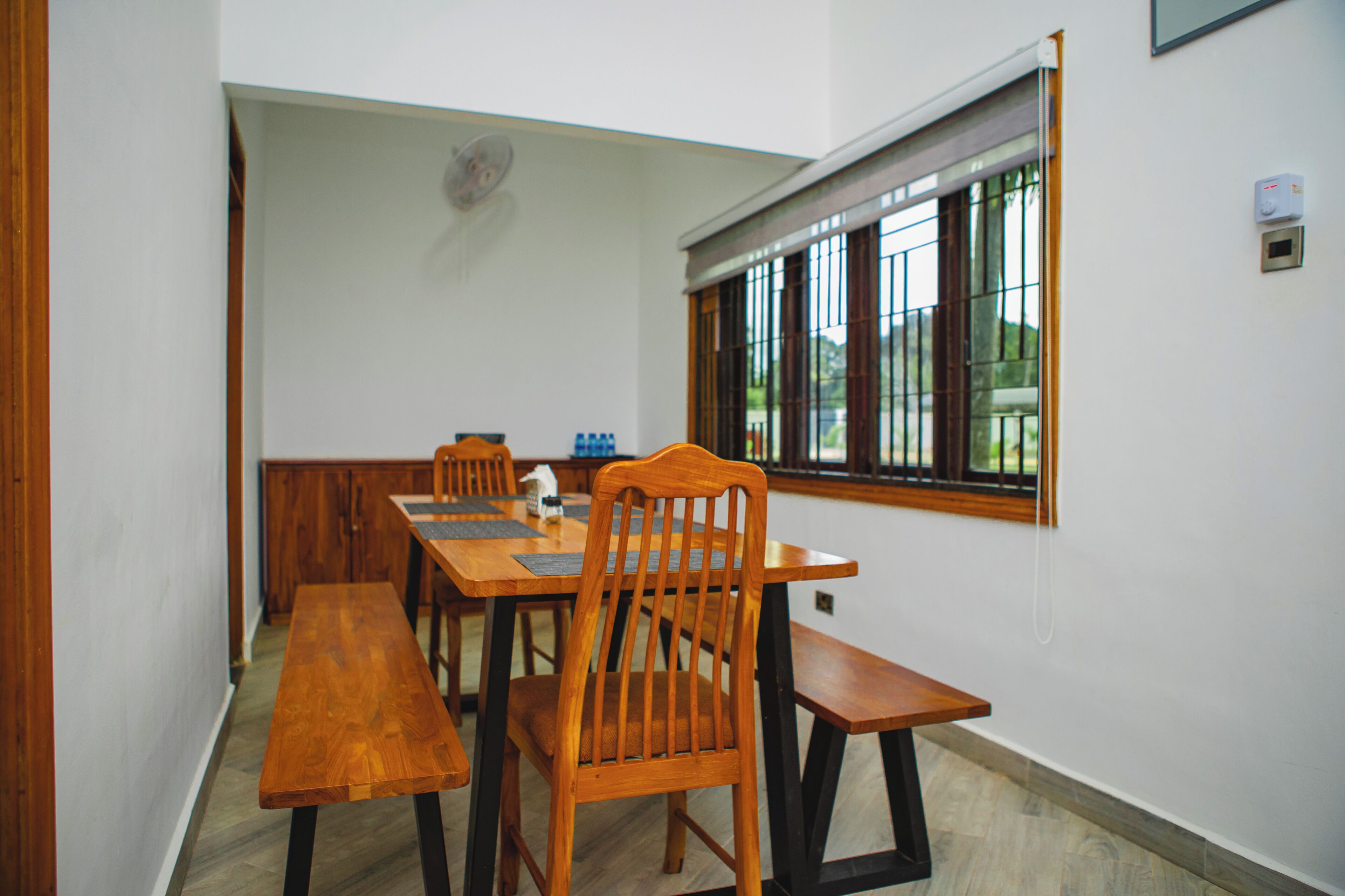 Ein Essbereich mit Holztischen und -stühlen, einem Fenster mit Gitterrost und einem Wandventilator.