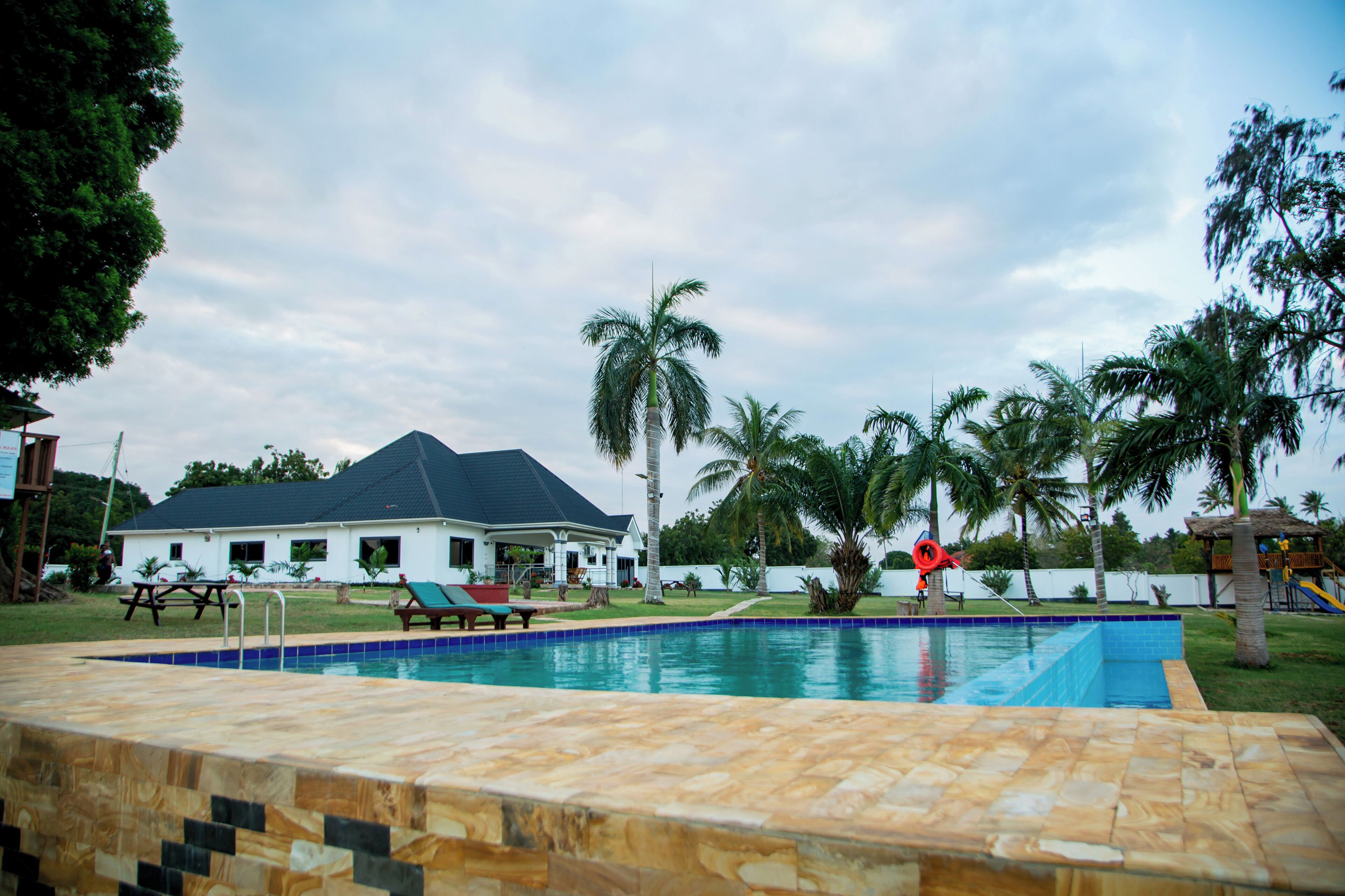 Una piscina con solárium empedrado, palmeras y una casa al fondo.