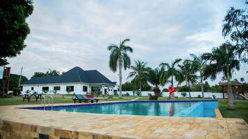 Una piscina con solárium empedrado, palmeras y una casa al fondo.