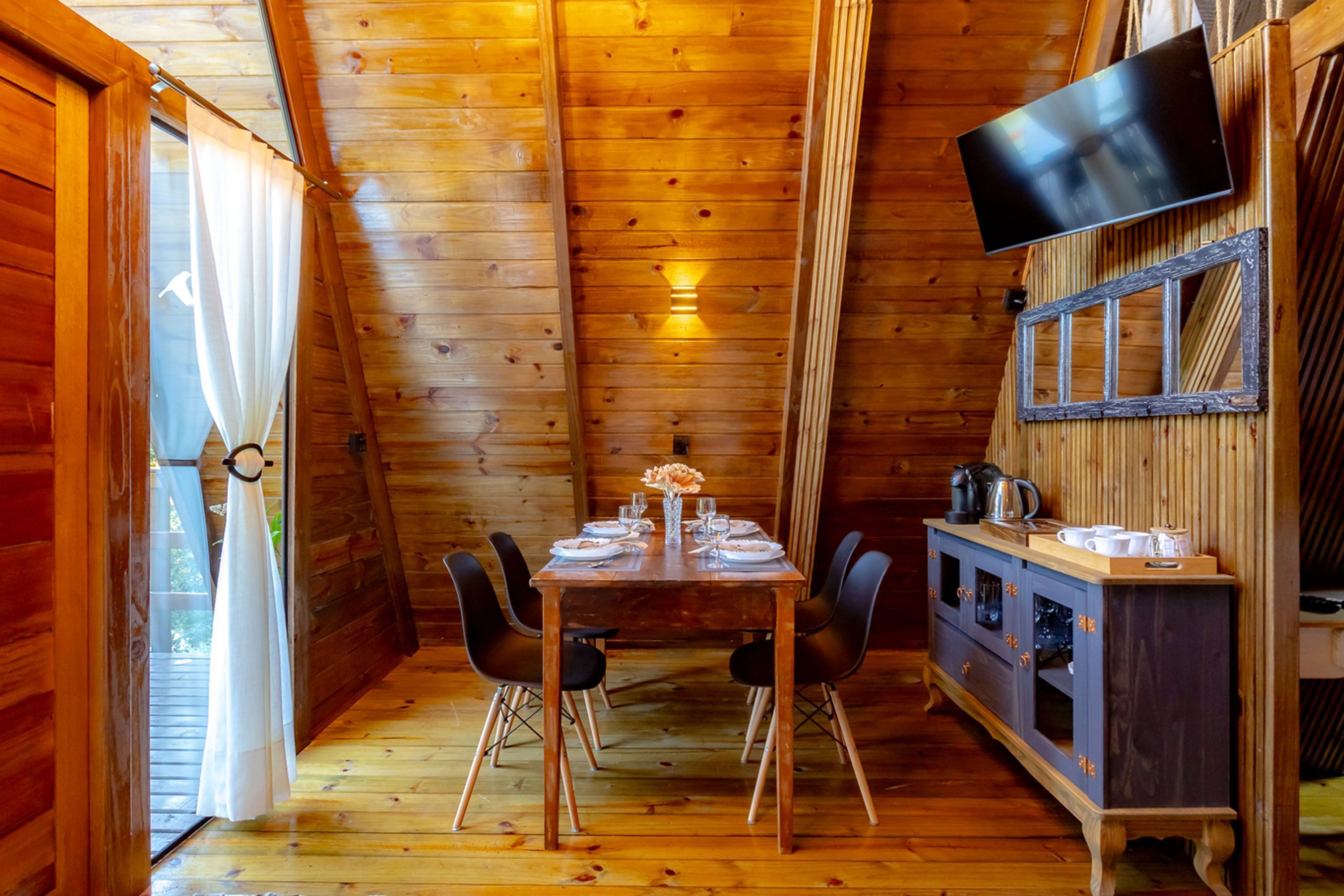 Interior de una cabaña de madera con una mesa de comedor preparada para cuatro personas, un televisor montado en la pared y un armario con utensilios de cocina.