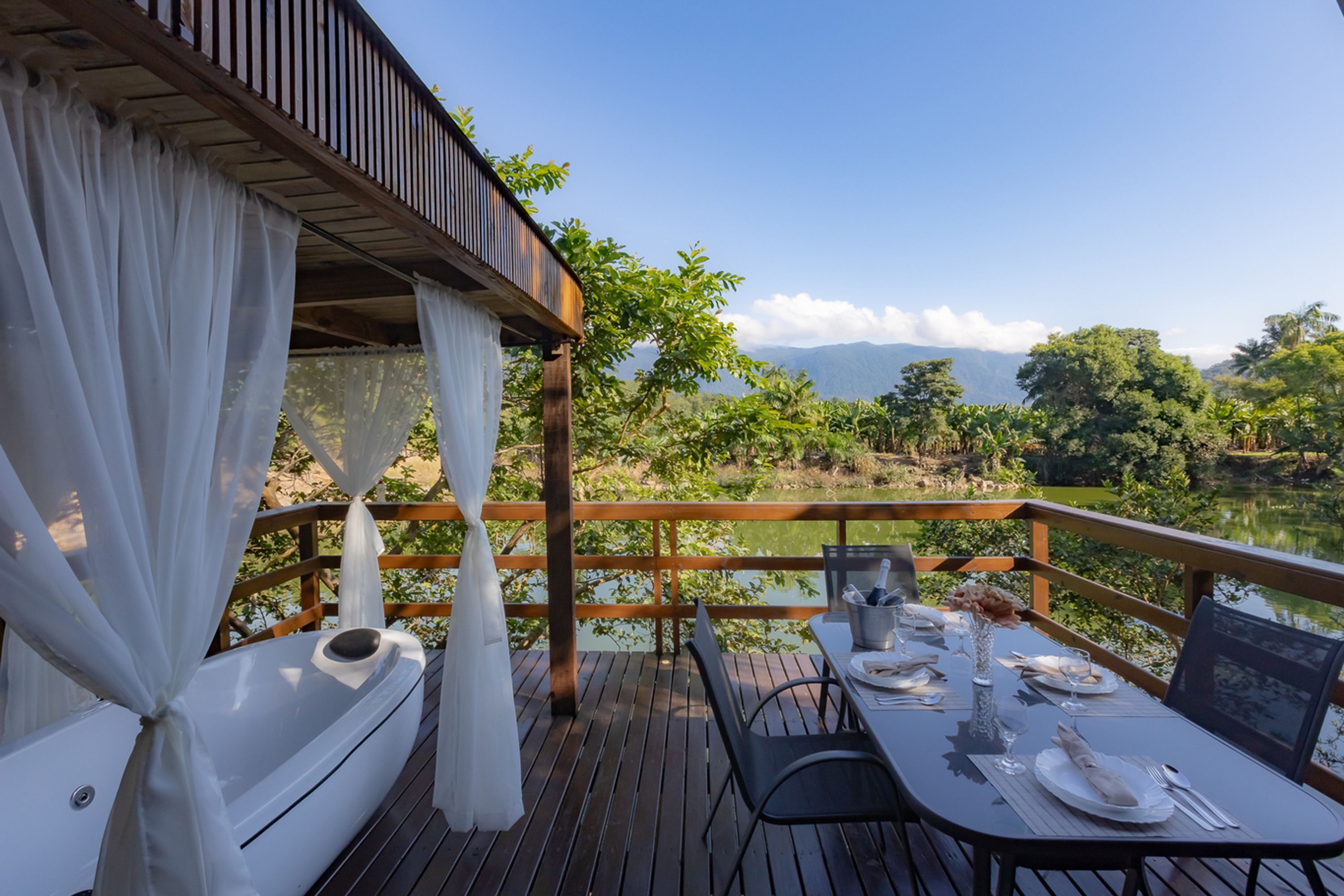 Um deck de madeira com uma mesa posta para refeições, com vista para um lago e montanhas.