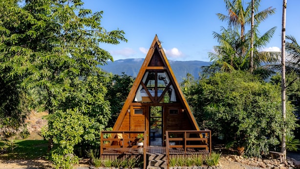 Una baita a forma di A in legno con fronte in vetro, immersa nel verde rigoglioso e con montagne sullo sfondo.