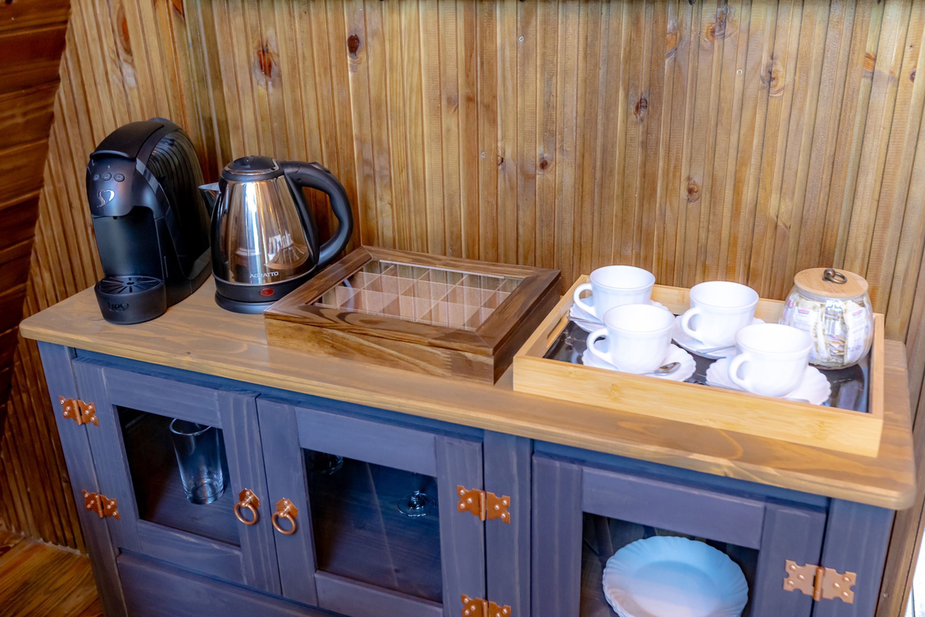 A wooden cabinet with a coffee maker, a kettle, and a tray of white cups and saucers.