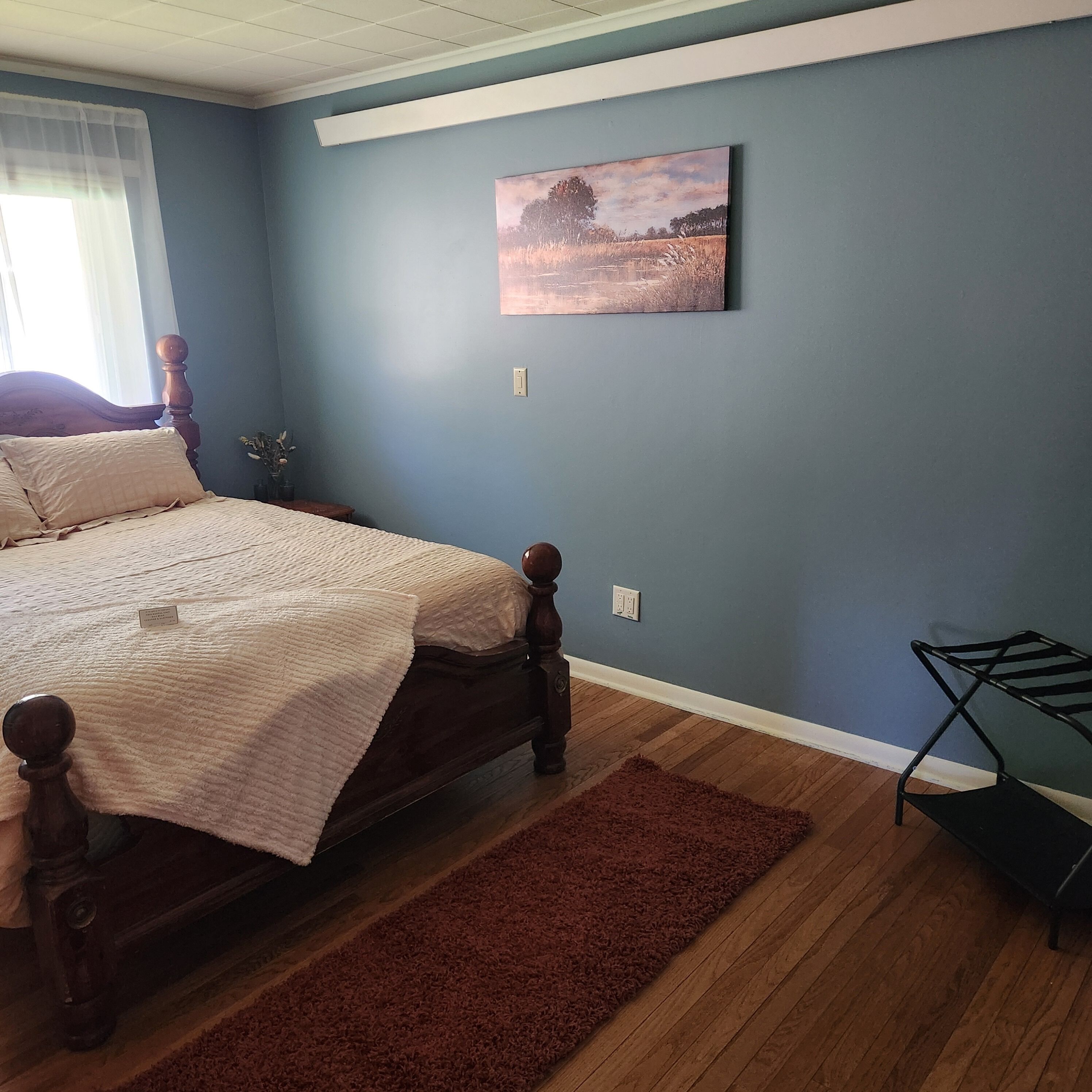 A bedroom with a bed, a nightstand, a wall-mounted painting, and a small table with a plant.