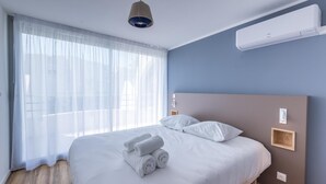 A modern hotel room with a large bed, white linens, a wooden headboard, and a large window with sheer curtains.