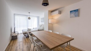 A modern hotel room with a wooden dining table, white chairs, a sofa, a TV, and a large window with sheer curtains.