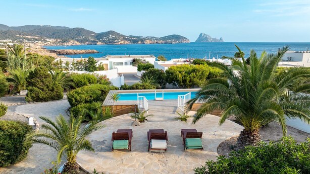 Un área de piscina con sillones reclinables, una terraza de madera y vistas al mar y las montañas.
