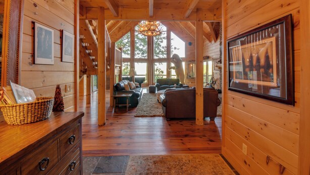 A wooden cabin interior with a wooden floor, a fireplace, a sofa, and a large window.