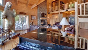 A log cabin interior with a stone fireplace, a bear statue, a glass-top table, and a second-floor balcony.