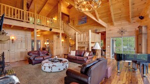A spacious living area with a grand piano, leather sofas, and a high wooden ceiling with a chandelier.