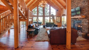 A spacious log cabin interior with a stone fireplace, wooden staircase, and a large window offering a view of the outdoors.