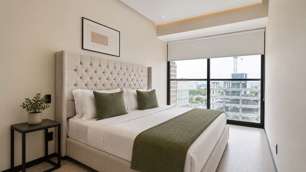 A modern bedroom with a tufted headboard, a large window with a city view, a bedside table with a plant, and a bed with a green blanket. - Bright 3BR Apartment | Minutes from Chapultepec (Guadalajara)