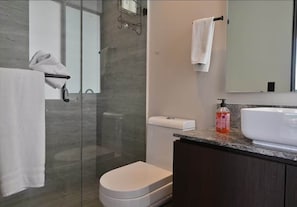 Un baño moderno con cabina de ducha de vidrio, inodoro blanco, lavamanos con encimera de mármol y espejo sobre el lavamanos.