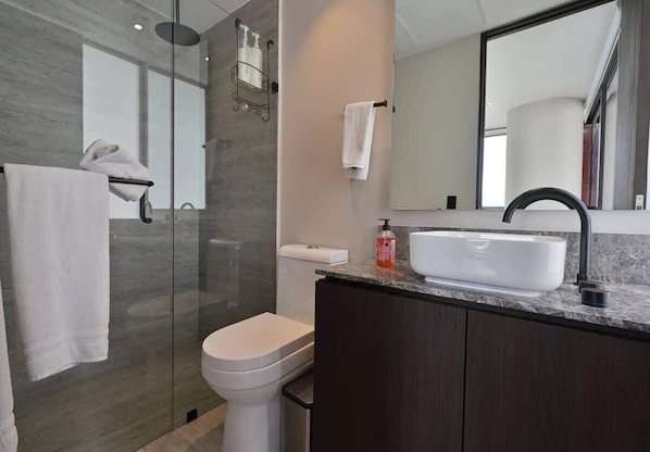 Un baño moderno con cabina de ducha de vidrio, inodoro blanco y tocador con lavamanos blanco y muebles oscuros.