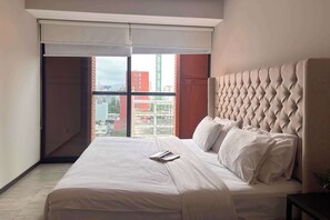 A hotel room with a large bed, a window with blinds, and a view of buildings.