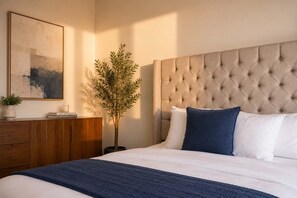 A bedroom with a tufted headboard, a bedside table with a plant, a framed picture, and a bedside lamp.