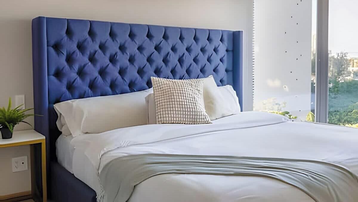 Ein ordentlich bezogenes Bett mit blauem Kopfteil, weißer Bettwäsche und einem karierten Kopfkissen.