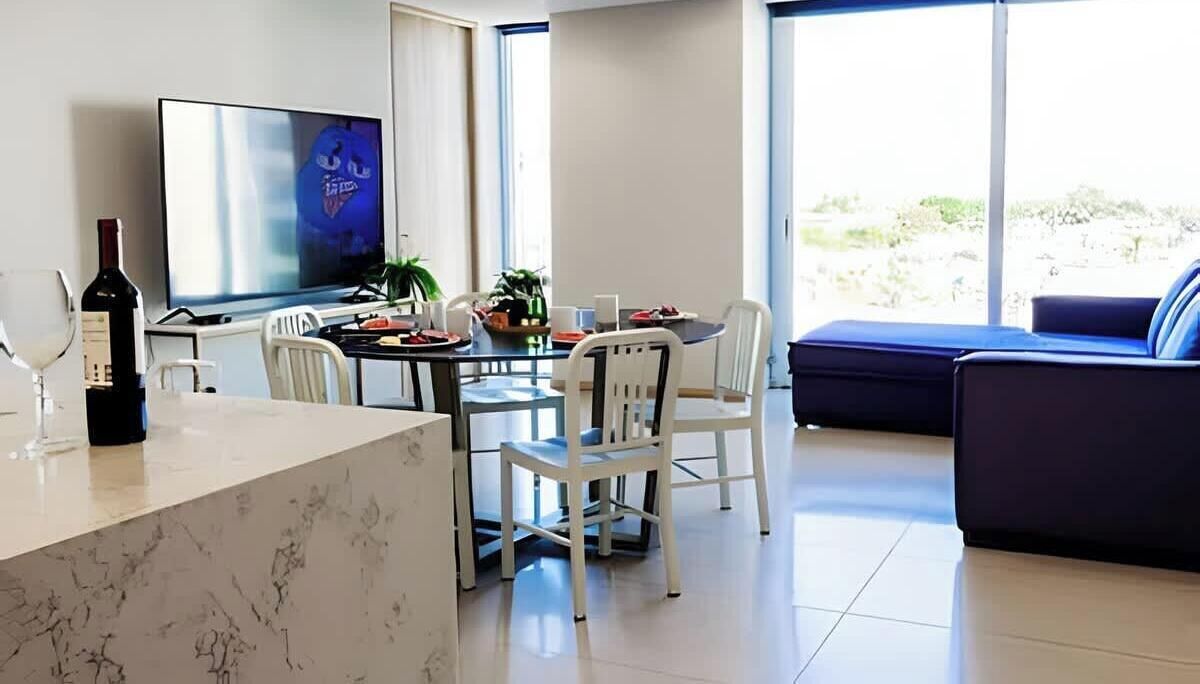 A modern living room with a dining area, a flat-screen TV, and a large window with a view of the outdoors.