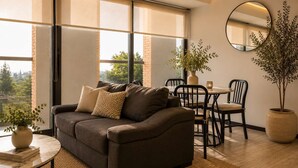 Ein modernes Wohnzimmer mit einer Couch, einem Esstisch, Stühlen, einer Topfpflanze und einem großen Fenster mit Jalousien.