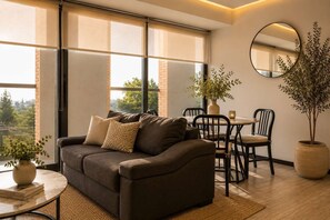 Ein modernes Wohnzimmer mit einer Couch, einem Esstisch, Stühlen, einer Topfpflanze und einem großen Fenster mit Jalousien.