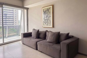 A modern living room with a grey sofa, a framed abstract artwork, and a large window offering a view of buildings and greenery. - Modern Apartment | Luxury, Comfort, and Location (Guadalajara)