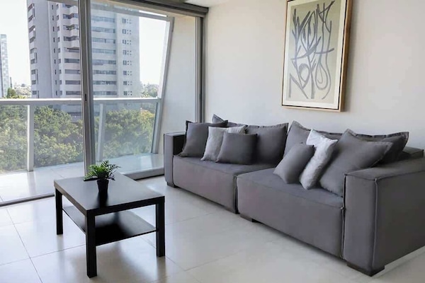Bright living room w/ plush sofa seating & floor-to-ceiling windows framing sweeping city views.