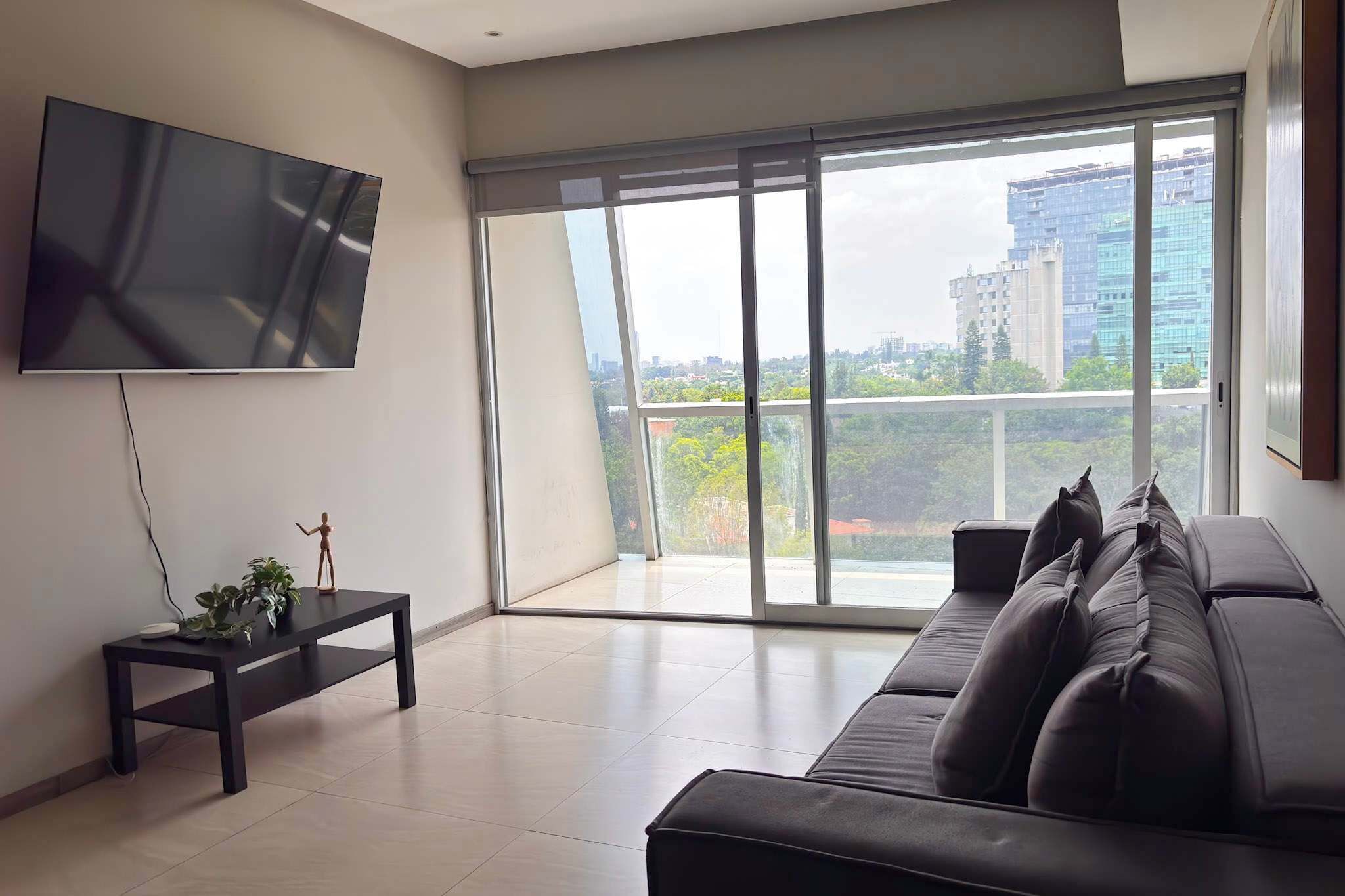 A modern living room with a large flat-screen TV mounted on the wall, a dark sofa, a small table with a plant, and a view of a cityscape through large windows.