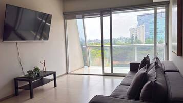 A modern living room with a large flat-screen TV mounted on the wall, a dark sofa, a small table with a plant, and a view of a cityscape through large windows.