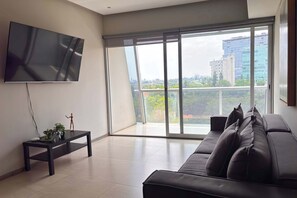 A modern living room with a large flat-screen TV mounted on the wall, a dark sofa, a small table with a plant, and a view of a cityscape through large windows. - Modern Apartment | Luxury, Comfort, and Location (Guadalajara)