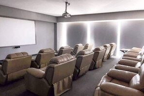 A room with rows of leather recliner chairs, a ceiling-mounted projector, and a blank screen on the wall.