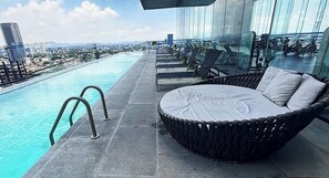 Een zwembad op het dak met een glazen balustrade, ligbedden en uitzicht op de stad.