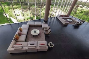 Un vaste espace de vie comprenant un grand canapé d’angle, une table basse et une vue sur un jardin luxuriant grâce à de grandes fenêtres.