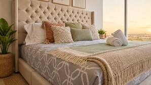 A bedroom with a tufted headboard, multiple pillows, a bedspread with a geometric pattern, a macrame throw, and a view of the city through a large window.