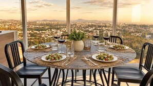 A round dining table with plates of food, glasses of wine, and a potted plant, set against a cityscape view.